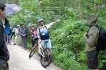 Eine Gruppe Menschen steht auf einem Waldweg. Ein Mann mit einem Mountainbike und Helm zeigt in die Ferne und spricht.